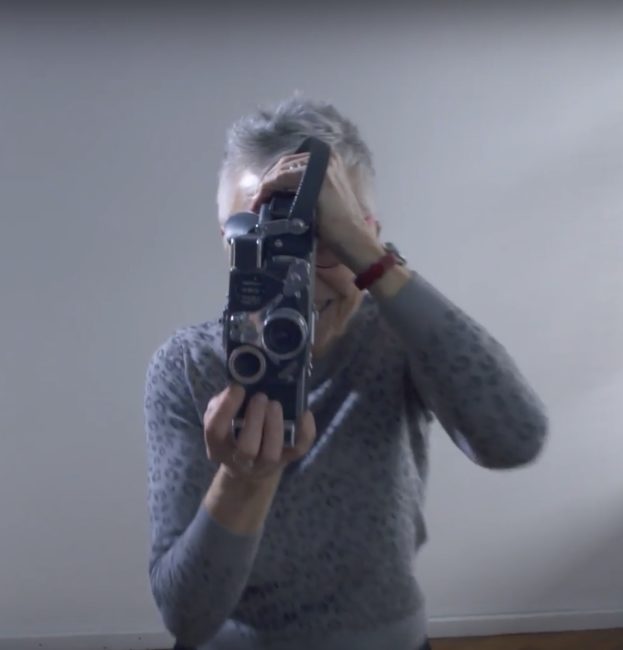 Medium frontal shot of the filmmaker holding the Bolex in both hands. Her right hand is on the underside of the Bolex, index finger on the release button, and her left hand is holding the top of the camera. Her eye is glued to the viewfinder.