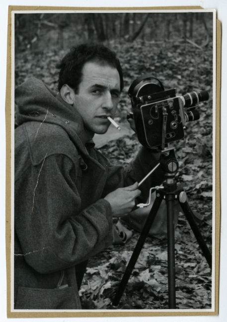 A young man looks at the camera that is photographing him. He is filming outdoors. He is standing behind his Bolex with a cigarette in his mouth, adjusting the frame. His right hand is on the handle of the tripod, guiding the camera. His left hand is next to the lenses.
