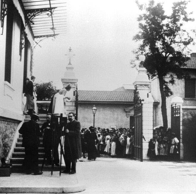 Two operators are standing next to their Cinématographe, outdoors in front of a large staircase. In the background, a crowd is gathered in front of the entrance to the property.