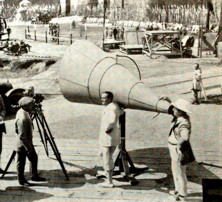 On a film set,  left foreground, the camera operator stands in front of a Bell & Howell, which is mounted on a tripod. On the right, the director stands in front of a huge megaphone. The sets, the extras and the rest of the film crew are visible in the background.