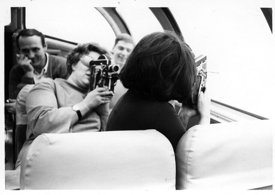 In a bus. A woman on the left, Wendy, films a woman, Joyce, whose back is to the photographer. Joyce films the outdoors through the bus window. Two men in the background smile as they watch.