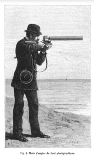 A man standing by the sea aims his gun at something out of sight. Slung across his shoulder is a box that protects the sensitive photographic plates from light.