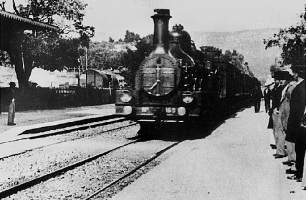 A train pulls into the station. Travellers on the right-hand side of the frame wait on the platform.