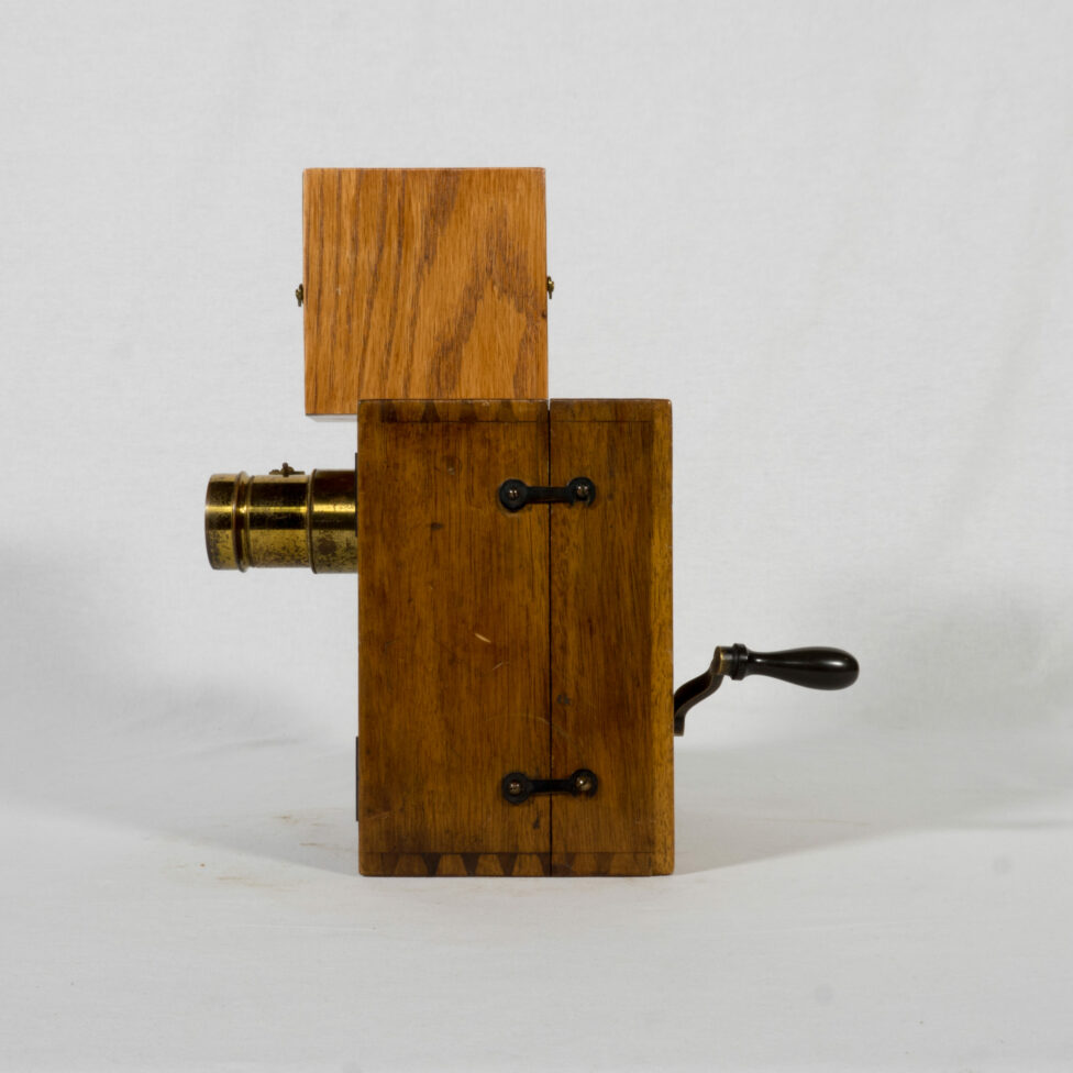 We can see the square body of the wooden camera topped by a smaller box that contains the blank film. The lens is visible at the front and the crank at the back. Two small hooks are positioned on the side and allow to maintain the camera closed.