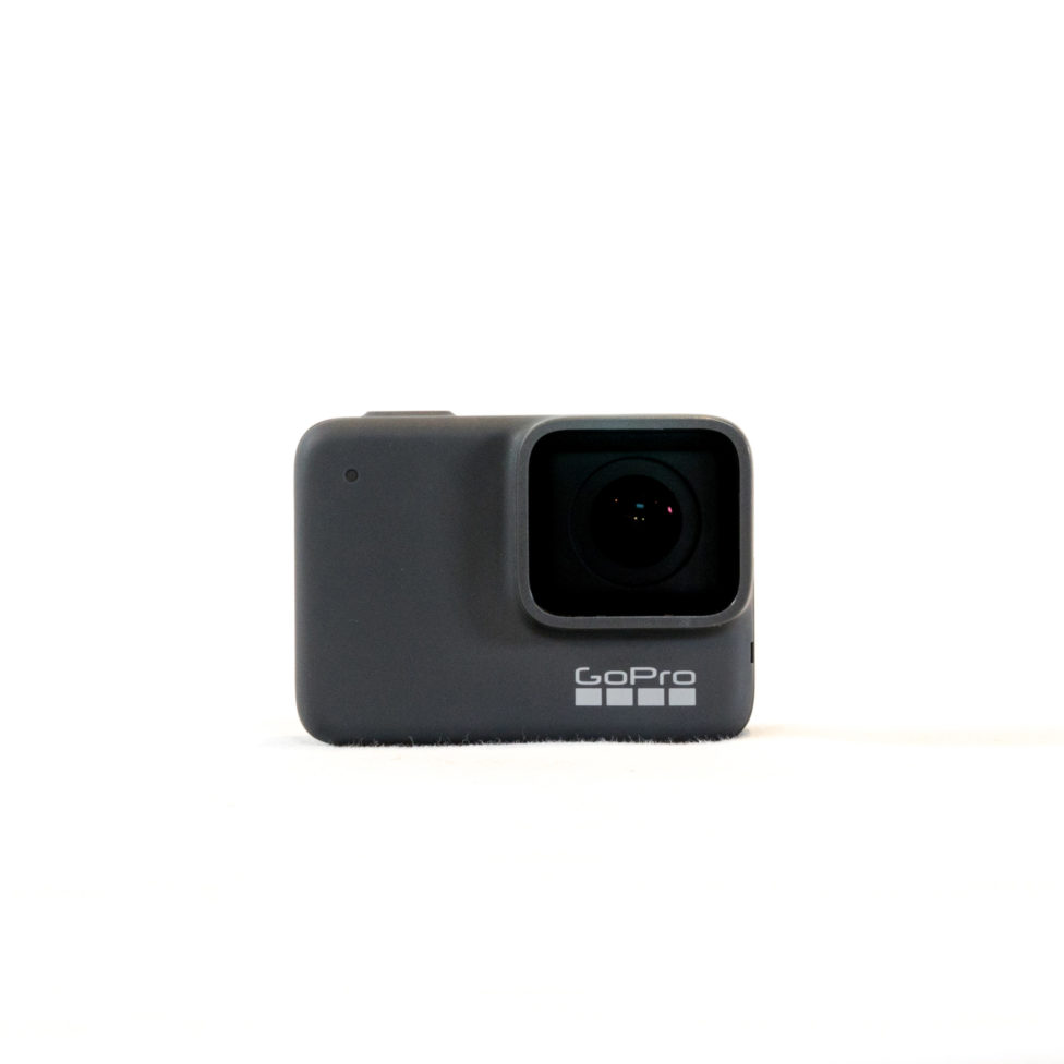 Photograph of the front of a small grey camera. A small square in the upper right of the camera contains the lens. Under the square, the company name, “GoPro,” and the four squares of its logo, all in white, are visible.