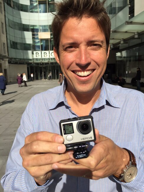 A smiling man stands in front of a BBC building. He is showing a GoPro camera to the photographer.