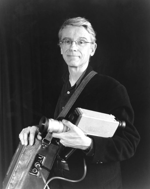 A man holds a Portapak camera on his left arm. The recorder is in a leather bag slung across his chest. He is posing with a smile.