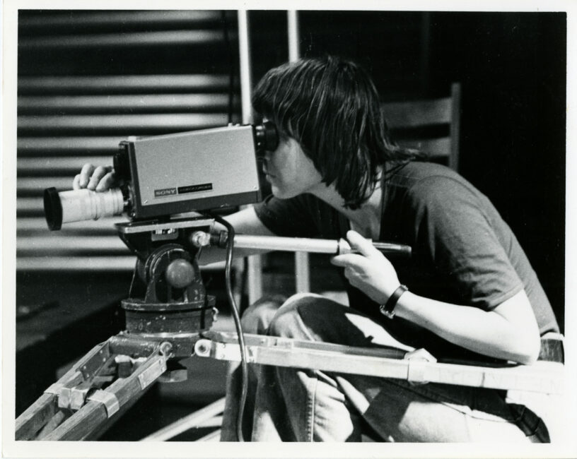 A woman is looking through the Portapak viewfinder. Her left hand is on the handle of the tripod, to guide the camera. Her right hand is adjusting the focus.