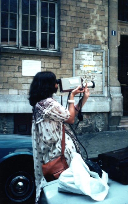 Nicole is holding the camera without using a tripod. Her eye is glued to the viewfinder, her right hand is supporting the underside of the camera and her left hand is on the zoom lens. The recorder is resting on a car beside her.
