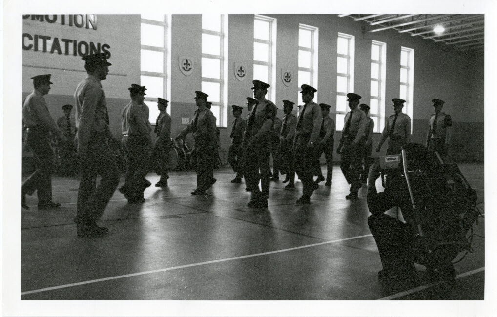 Police cadets practice marching in line in a large drill hall. The film director, holding the camera and the recorder, is on the right.