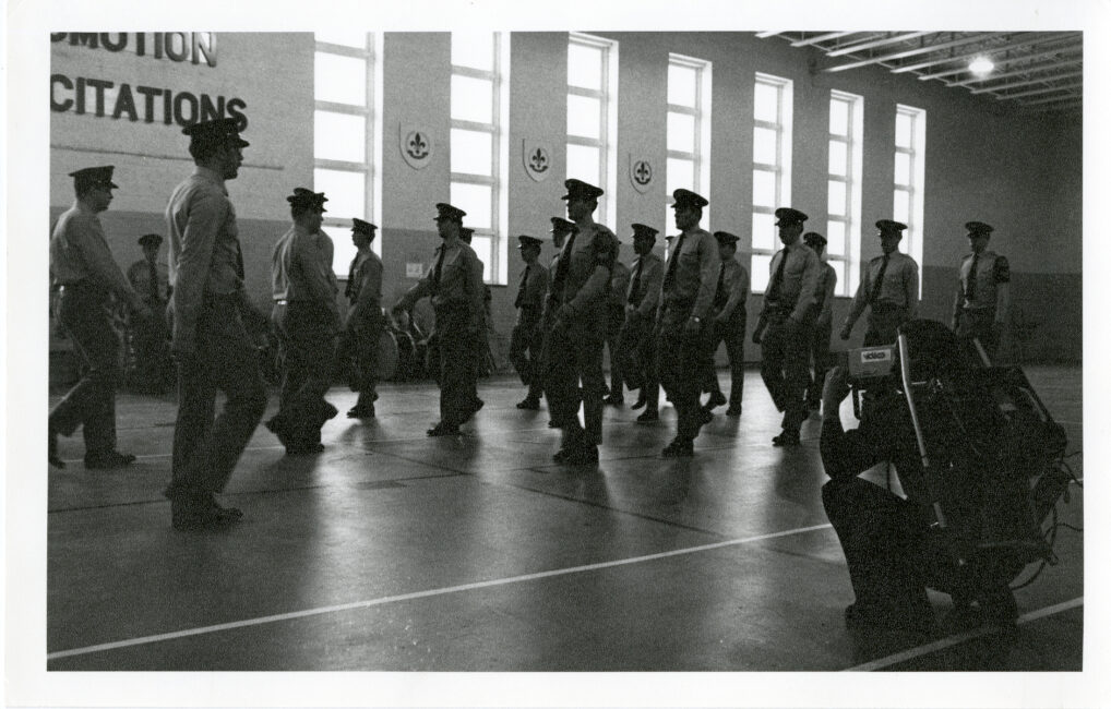 Les aspirants policiers s’entraînent à marcher en ligne dans leur grande salle de répétition. À droite, accroupi, se trouve le réalisateur avec la caméra et l’enregistreur.