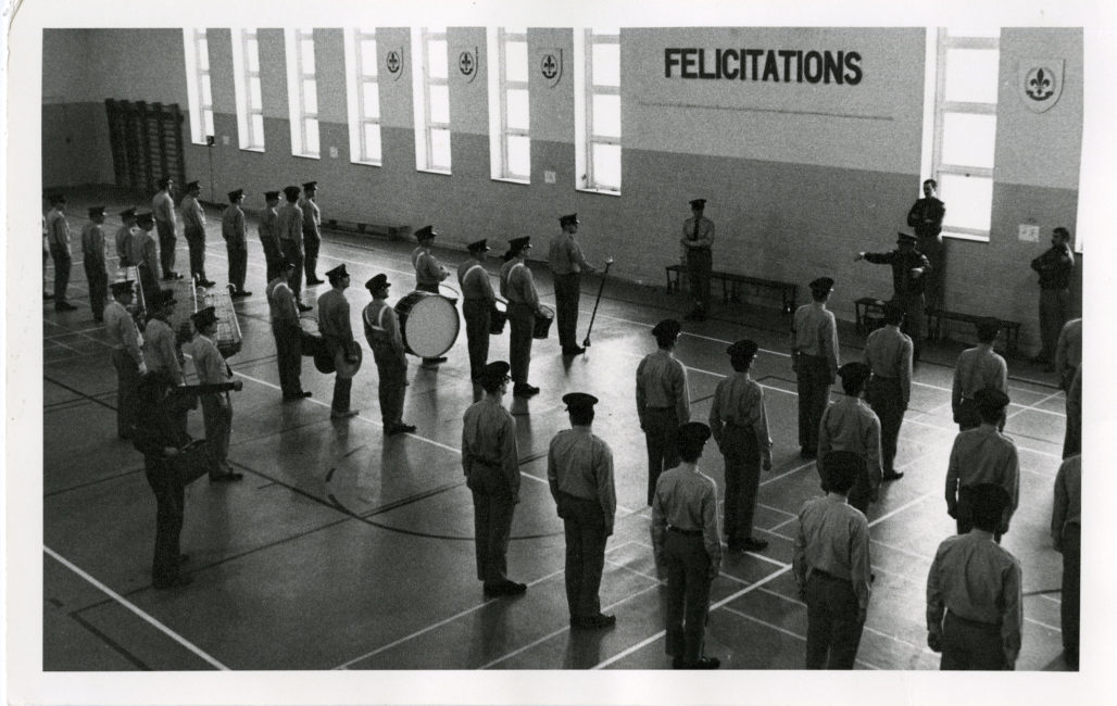 Police cadets are lined up in a large room, facing the windows. They take up the entire room. On the bottom left, the director stands holding the camera and the recorder.