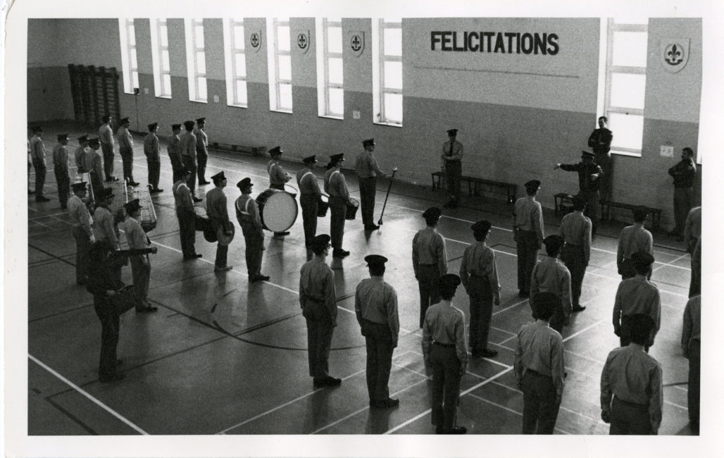 Les aspirants policiers sont alignés et tous orientés vers les fenêtres de la grande pièce. Ils occupent tout l’espace. En bas à gauche de la photographie se trouve le réalisateur équipé de la caméra et de l’enregistreur.