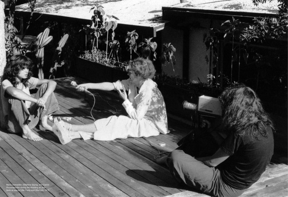 Three women are seated. The one on the left is being interviewed. To the right of her, another woman holds out a microphone to record her words. She is supporting her right arm with her left hand. The woman on the right of the photo is filming them. Her backed is rounded and she appears to be distancing herself from her subjects.