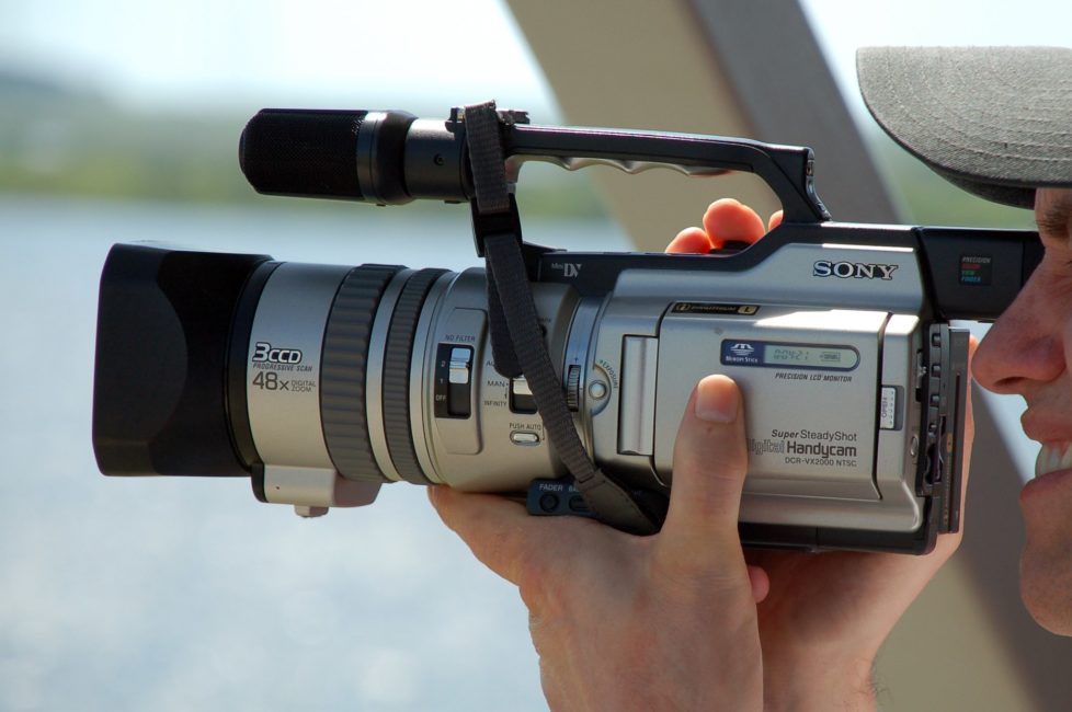 A man is holding a small Sony camera. With his eye on the viewfinder, he holds the camera in his two hands. The camera is approximately one and a half times the length of his hands.