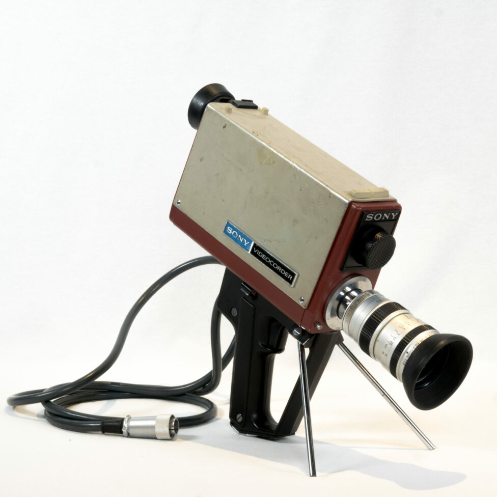The right side of the camera is visible. The camera is supported by its metal stand and its handle. The zoom lens is on the right, with the integrated microphone above it. The rectangular body of the camera is visible, as are the viewfinder and the cable on the left.