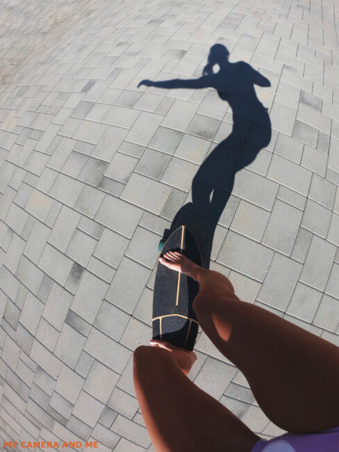 A person rides a skateboard. She is holding a GoPro in her right hand. Her legs and shadow can be seen.