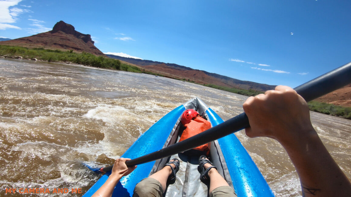 The front end of a kayak is visible, as are the kayaker’s arms and legs. She is in the middle of a brown river. A mountain can be seen in the distance.
