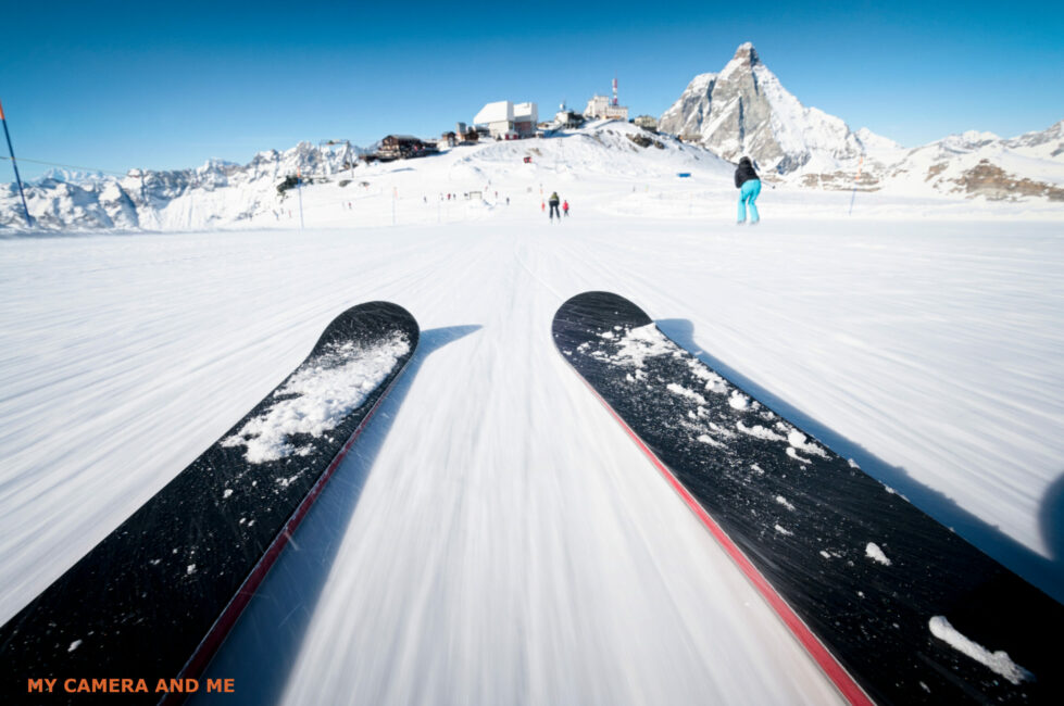 A pair of skis takes up three-quarters of the frame. The snow appears to be striated, giving the impression of fast movement. The camera is very close to the ground.