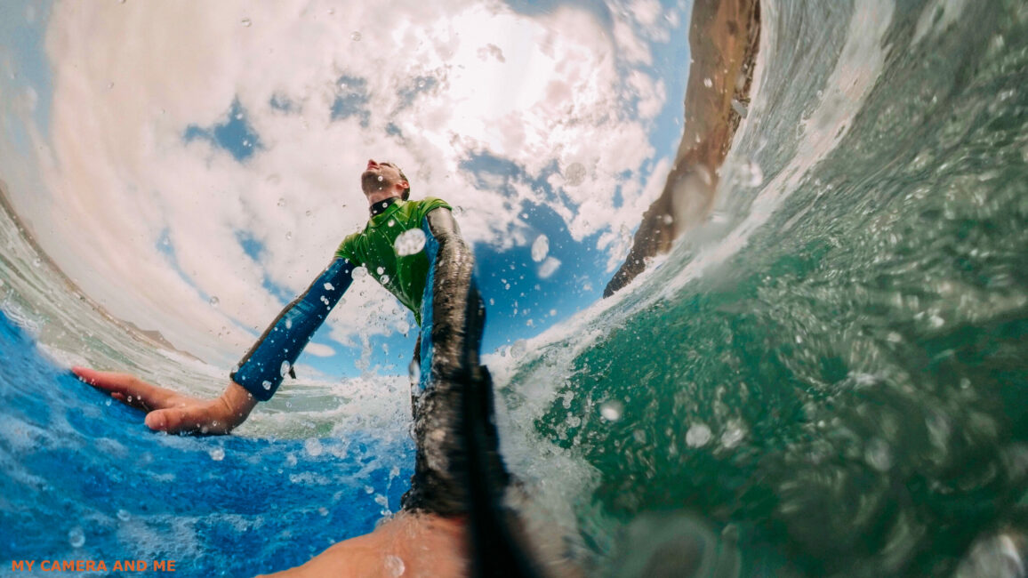 Low-angle photo of a man on a surfboard. The GoPro distorts the length of his arms.